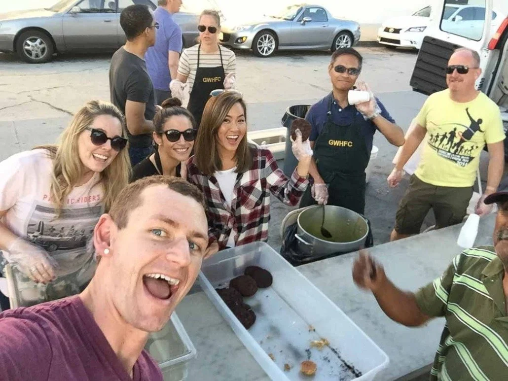 Group of people gathered outside around a table with baked goods, some wearing aprons, smiling and taking selfies, with cars parked in the background.