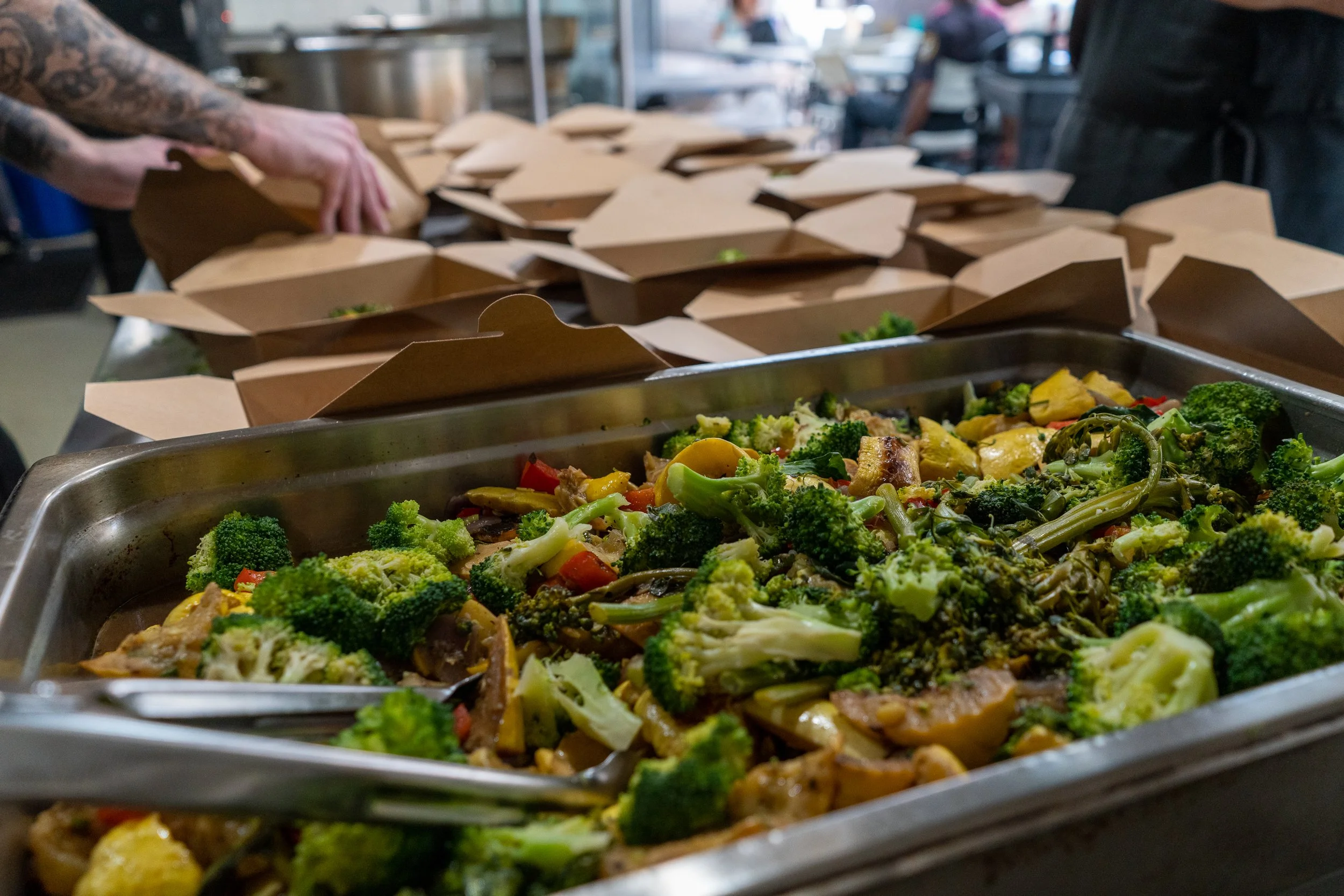 A tray of assorted cooked vegetables, including broccoli and bell peppers, on a buffet table with brown takeout containers and people in the background.