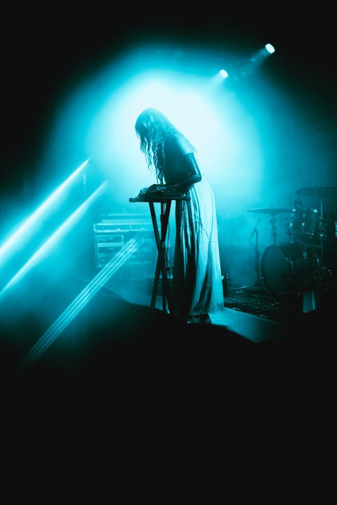 A woman standing on stage with long, wavy hair, wearing a long dress, illuminated by blue stage lighting, with a keyboard and drum set in the background, during a live music performance.