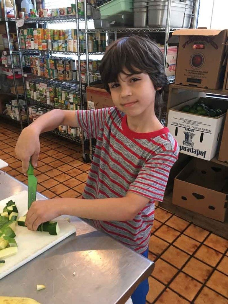 A young boy with brown hair, wearing a red and gray striped t-shirt, is slicing zucchini with a green knife on a white cutting board in a kitchen or store with shelves of canned goods in the background.