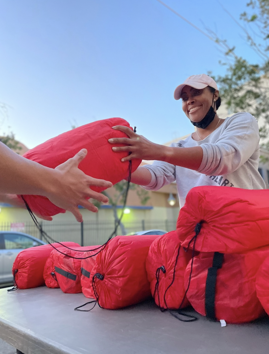 A woman wearing a light pink cap and a face mask under her chin is smiling and handing a red sleeping bag to another person. Several more red sleeping bags are laid out on a table outdoors, with trees, cars, and a building in the background.