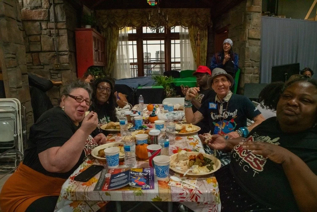 Group of people enjoying a meal at a festive gathering around a decorated table with plates of food, drinks, and fall-themed decorations in a cozy room with stone walls and a window.