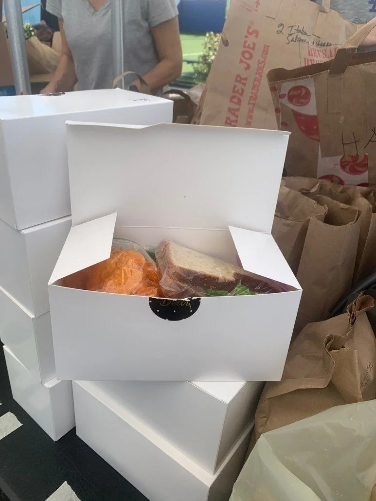 Open white takeout food box containing a sandwich and a wrapped item, placed on top of stacked white boxes, surrounded by brown paper bags, with people and a green outdoor area in the background.