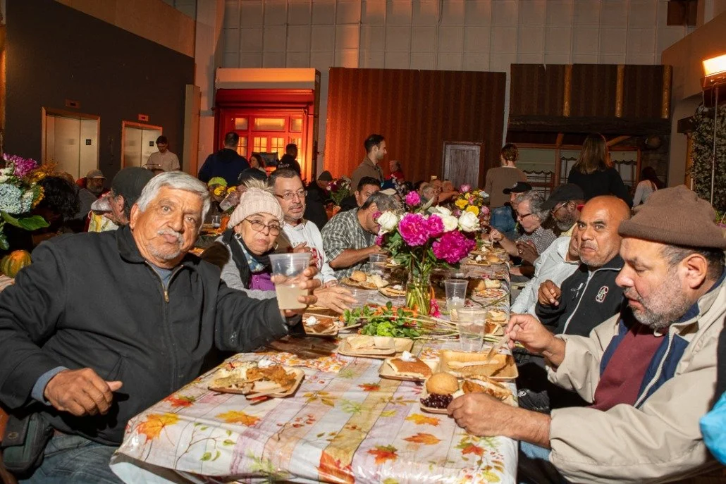 A diverse group of people gathered at a long dining table with fall-themed tablecloth and floral centerpiece, enjoying a meal together in a warmly lit room.