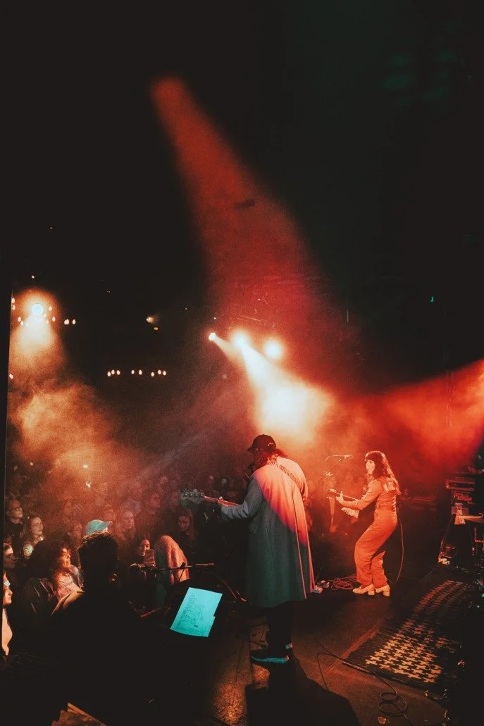 Musicians performing on stage with orange and yellow stage lights illuminating an audience in a dark concert venue.