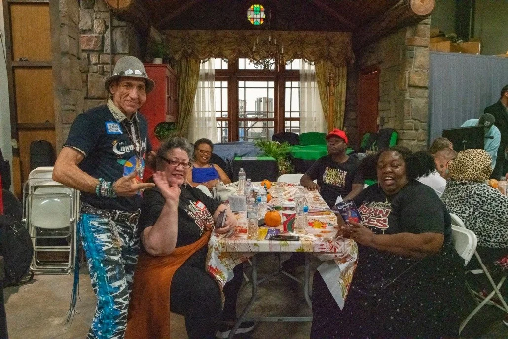 Group of people gathered around a decorated table in a rustic indoor setting, with some smiling and waving at the camera. A man stands to the left, wearing a colorful hat and patterned pants, while others are seated, engaging and smiling.