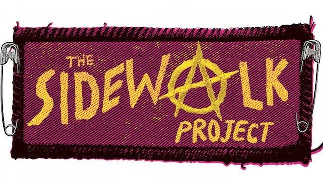 A fabric sign with frayed edges displaying the words 'The Sidewalk Project' in yellow, with an anarchy symbol in place of the letter 'A' in 'Sidewalk'.