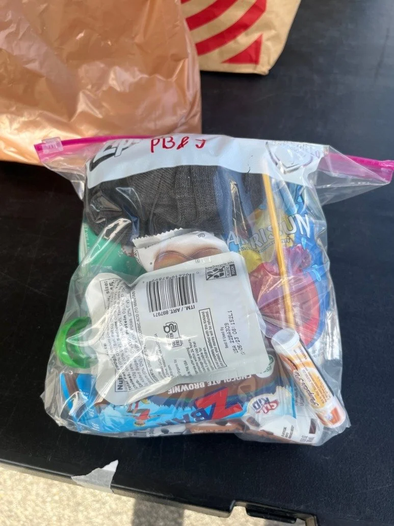 A clear plastic Ziploc bag containing various items, including a gray sock or glove, a tube of ointment or cosmetic product, a green and blue item, and a box of Ritz Zingers snack cakes. The bag is on a black surface, with a brown paper bag and a par