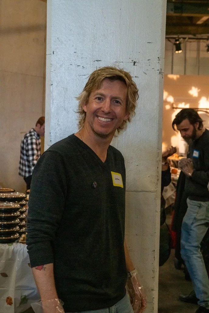 Smiling man with blonde hair wearing a black shirt with a yellow name tag, standing in a dimly lit indoor event space with other attendees and tables in the background.