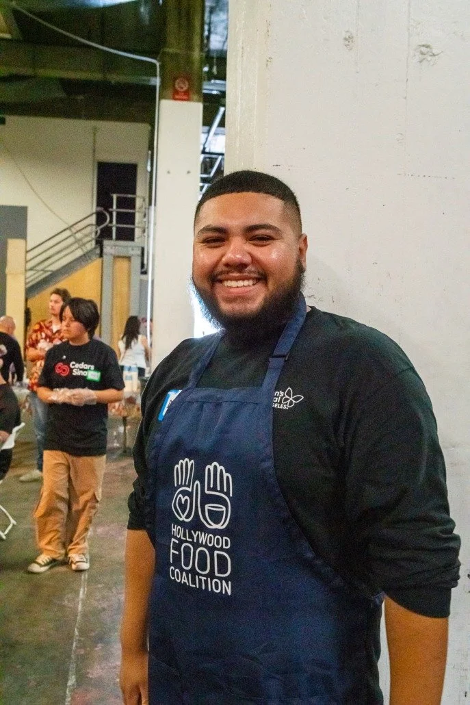 A smiling man in a black shirt and an apron with the Hollywood Food Coalition logo stands in an indoor setting, possibly a community event or food distribution.