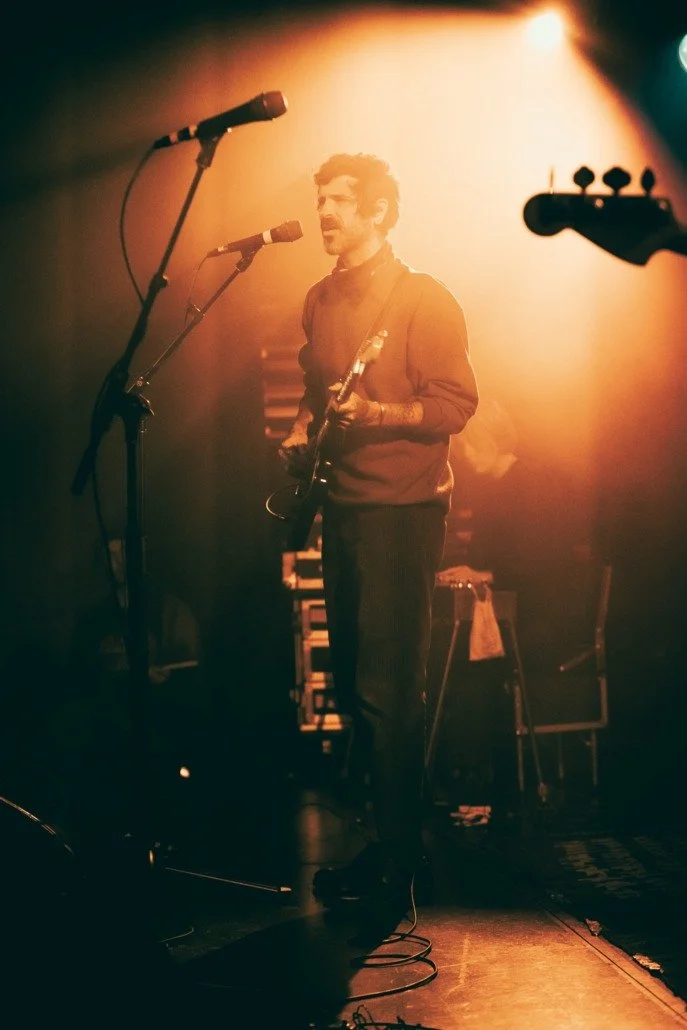 A man is on stage holding an electric guitar, singing into a microphone during a live music performance, with warm lighting creating a moody atmosphere.