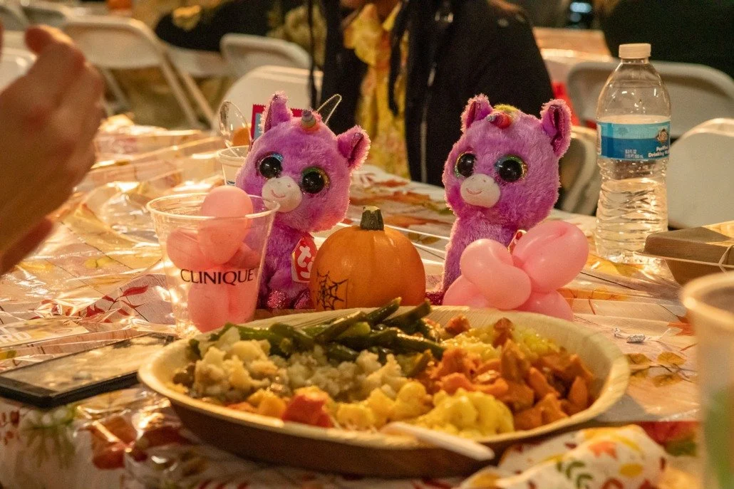 Decorative table setting with plush unicorn toys, pink balloons, a small pumpkin, a plate of food, a bottle of water, and a glass with pink objects, at a celebration or gathering.