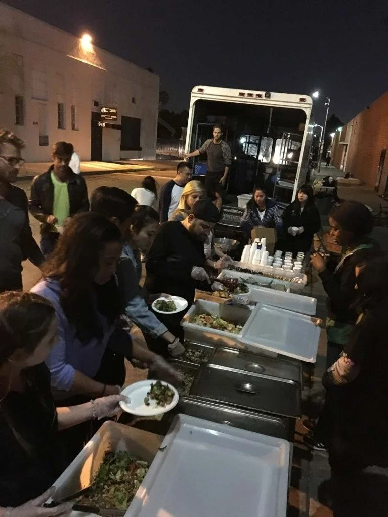 Community meal service with volunteers serving food outdoors at night, with a bus in the background.