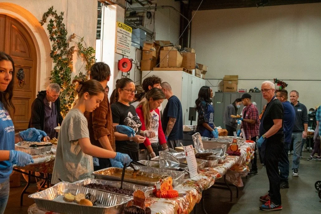 People serving food at a long table during a community meal event in a warehouse or large indoor space.
