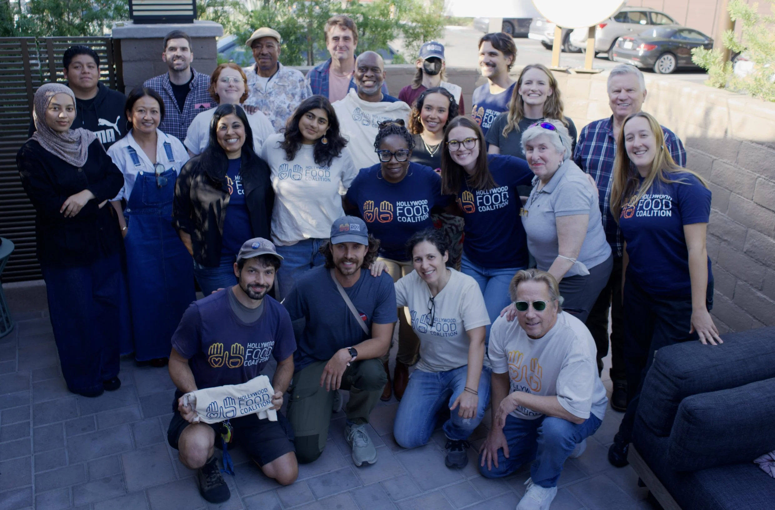 Group of diverse volunteers outdoors participating in a community event, some wearing hoodie and T-shirts with 'Hollywood Food Coalition' logo, smiling for the camera.