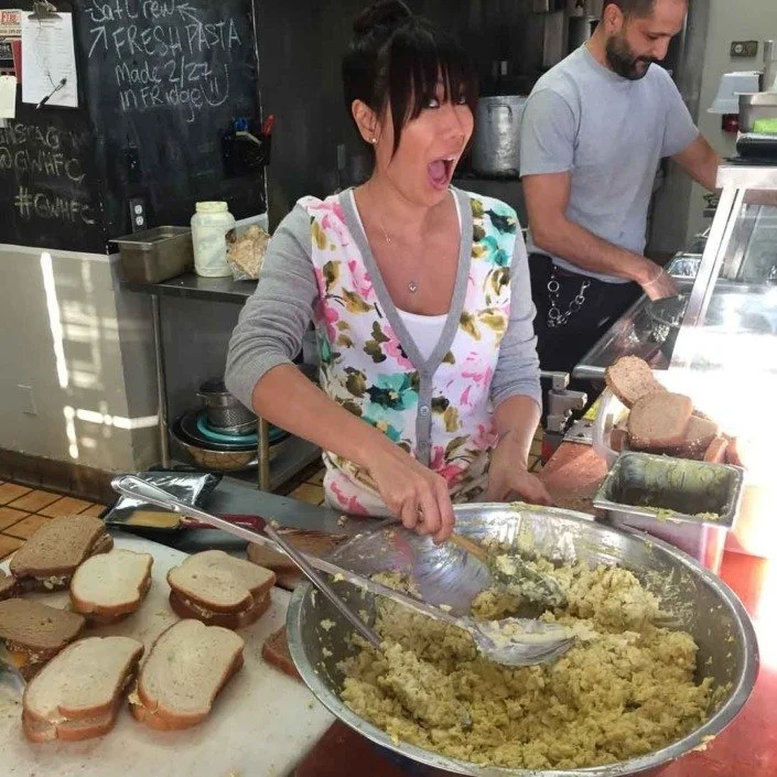 A woman wearing a colorful apron is standing in a kitchen, making a surprised or excited face while preparing mashed potatoes. She is holding a large spoon in a metal bowl filled with mashed potatoes. In front of her, there are slices of bread on a c