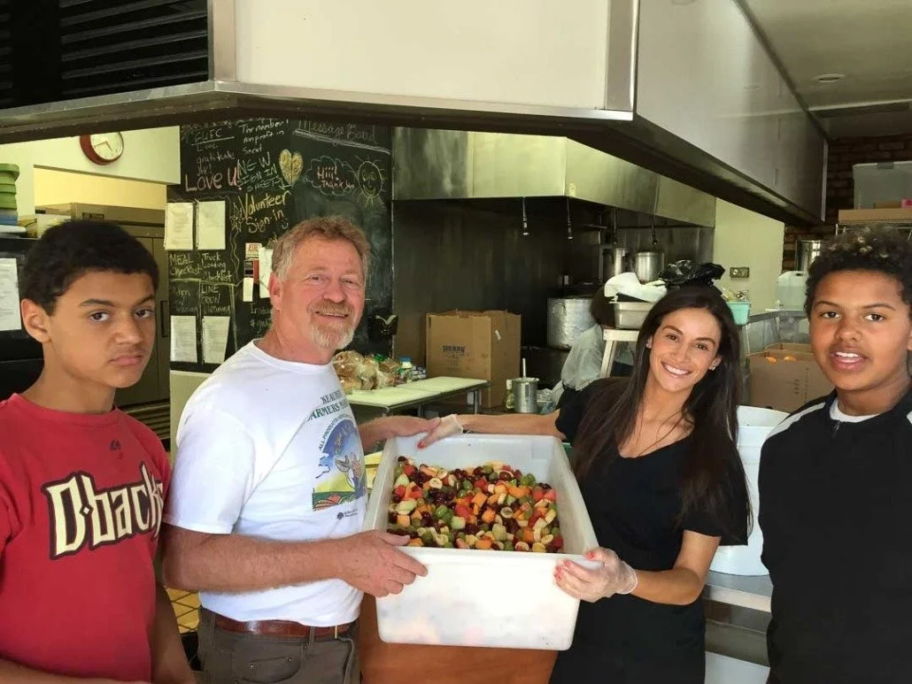 Four people in a kitchen holding a large tray of chopped vegetables and fruit.