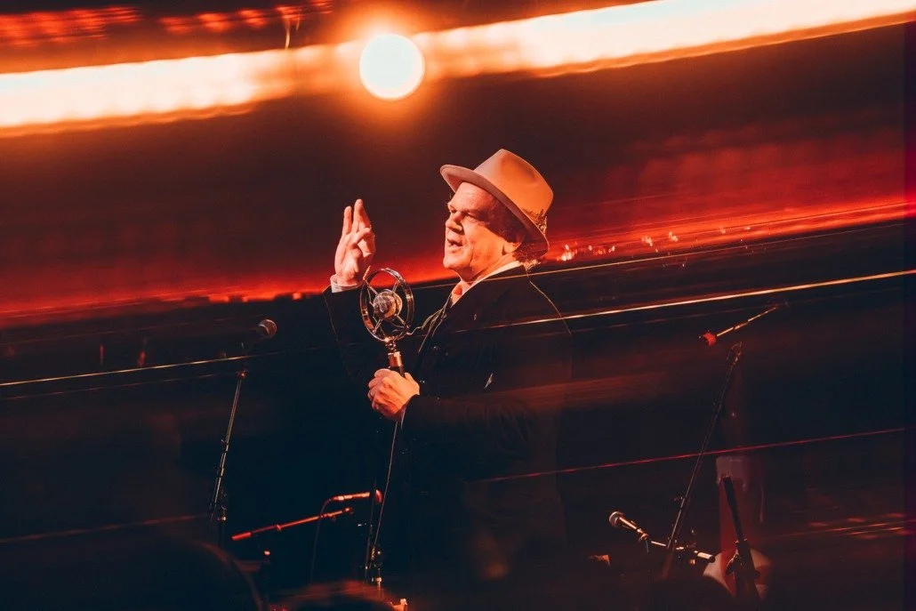 A man wearing a beige hat speaking into a microphone on a stage with a sunset and dramatic sky in the background.
