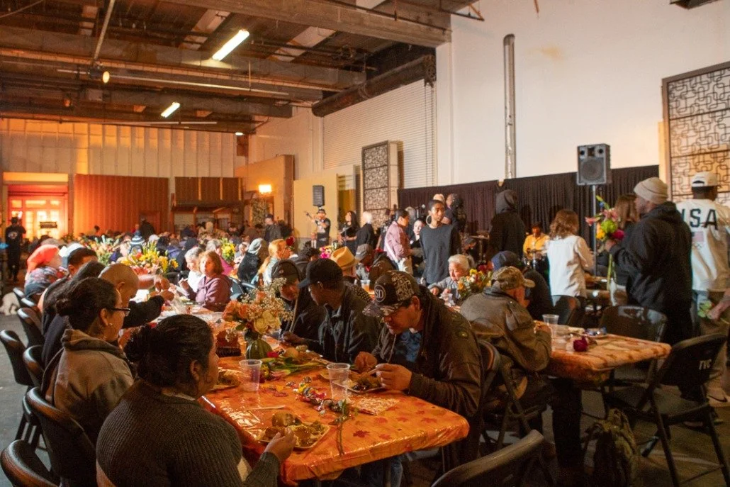 People gathered around long tables for a meal in a large indoor space with flowers and decorations on the tables.
