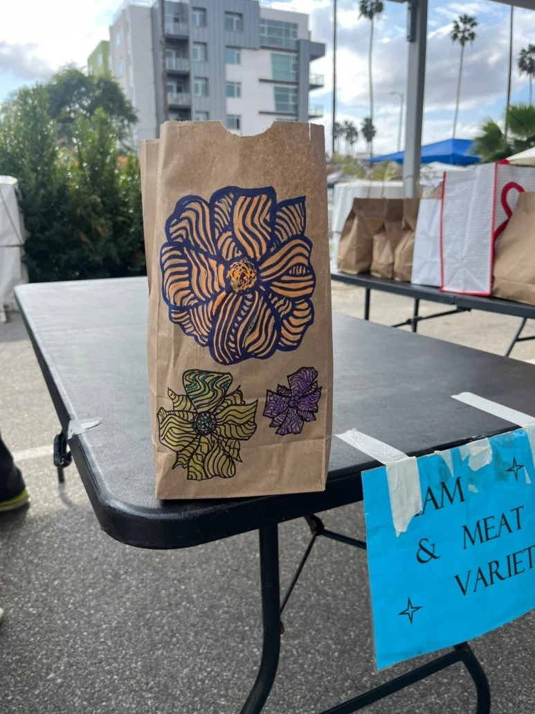 Brown paper bag with abstract floral designs in blue, green, purple, and yellow, sitting on a black table outdoors on a cloudy day.