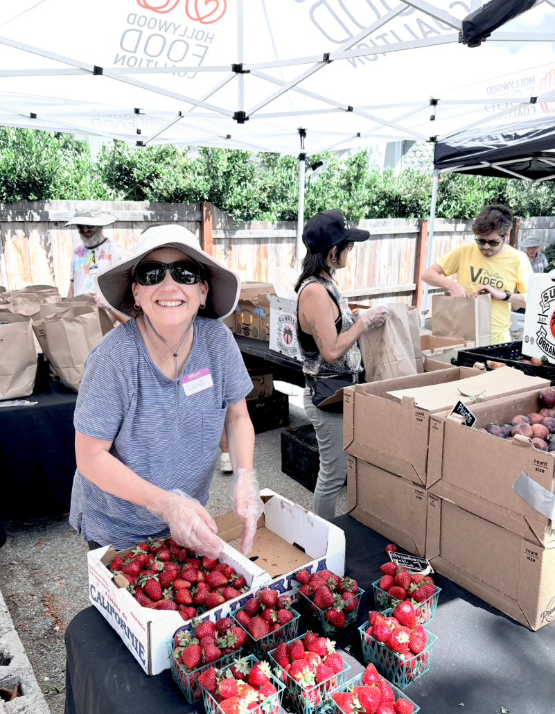 Smiling woman with sunglasses and wide-brimmed hat selling strawberries at an outdoor market stall, with other vendors and customers under tents in the background.