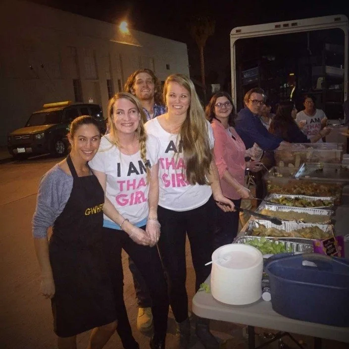 Group of people serving food at an outdoor event at night, with a food truck and parked cars in the background.