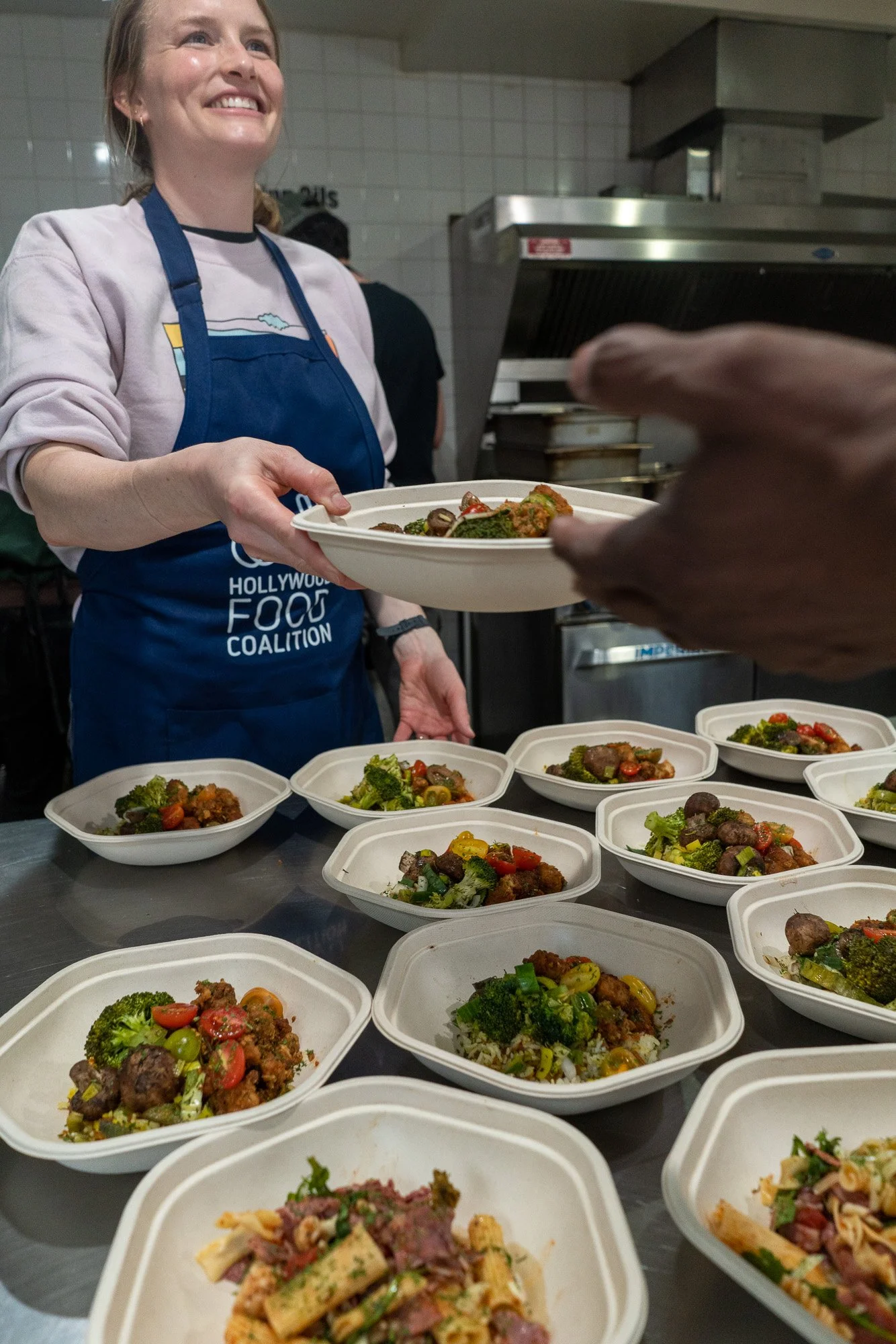 A woman in a blue apron with 'Hollywood Food Coalition' on it, smiling as she hands a bowl of food to someone. Several bowls of food, including pasta and vegetable dishes, are on a table in a kitchen setting.