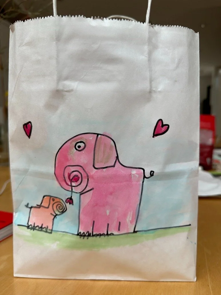 Hand-drawn illustration of two elephants, one larger pink elephant and a smaller tan elephant, with pink hearts around them, on a white paper bag.