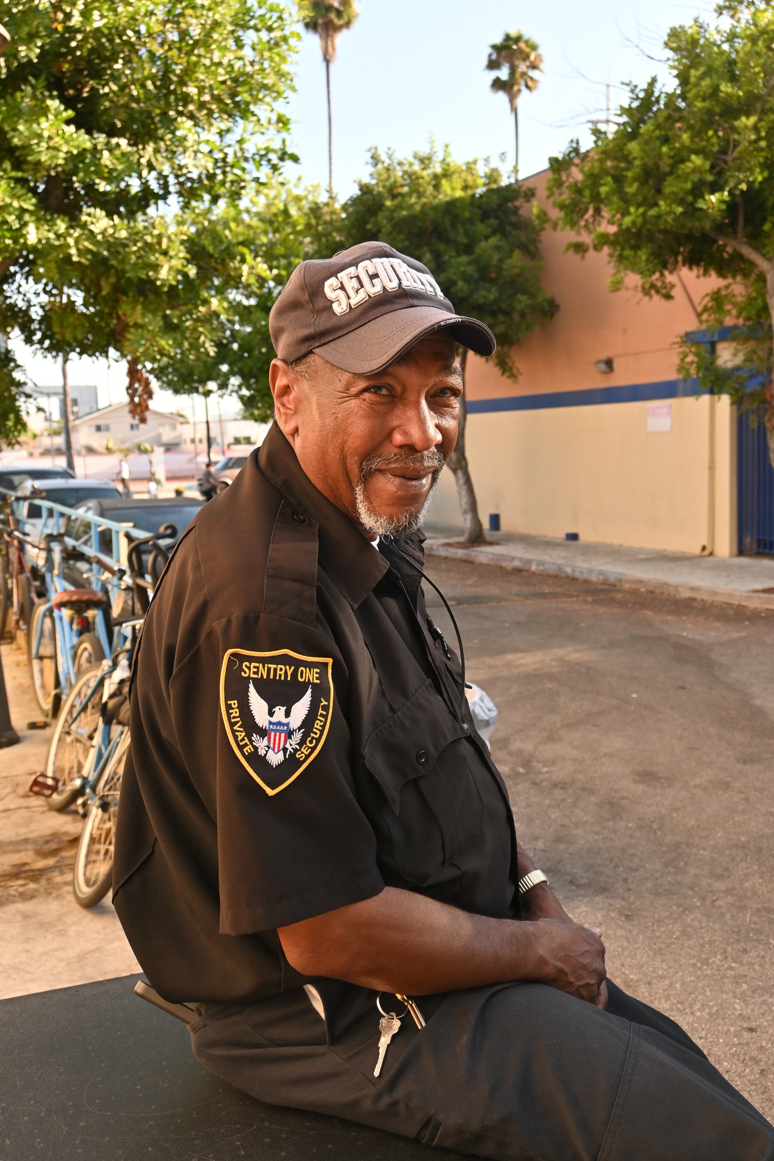 A security guard sitting outdoors near a row of bicycles, wearing a black uniform with a patch that reads 'Sentry One Private Security' and a cap labeled 'Security', smiling and looking at the camera.