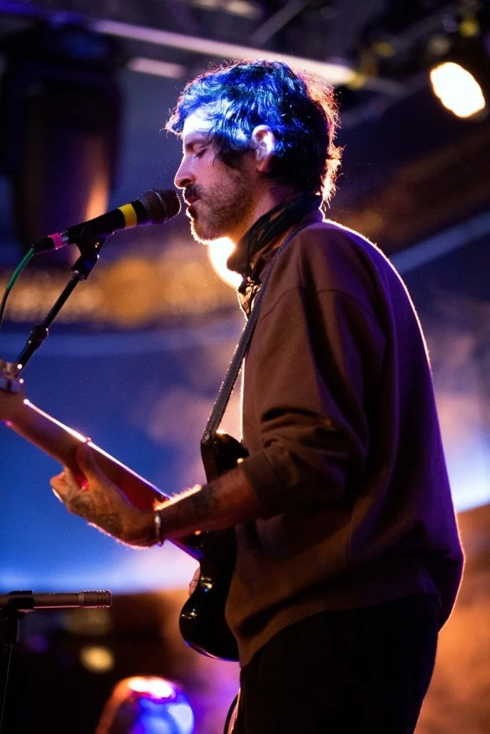 A male musician with dark, shaggy hair and a beard, singing into a microphone while playing an electric guitar on stage with colorful lighting in the background.
