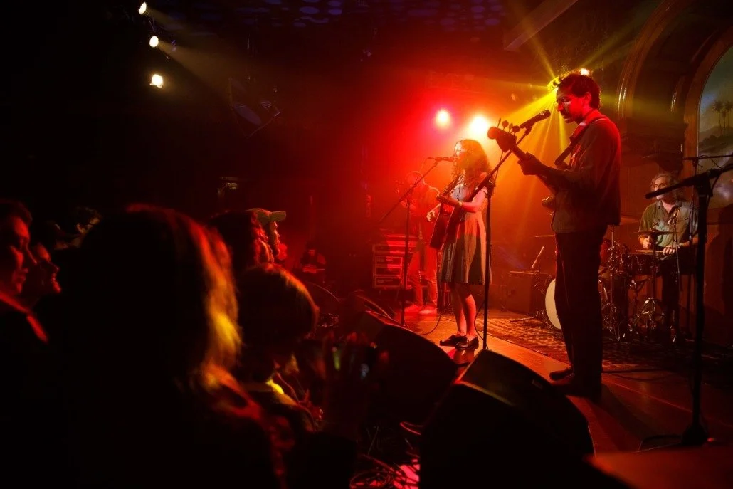 Musicians performing on stage with audience watching at a concert venue, illuminated by colorful stage lights.