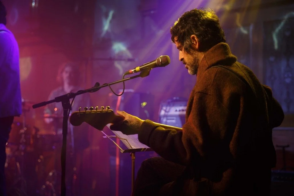 A man playing an acoustic guitar and singing into a microphone on stage during a live music performance, with vibrant stage lighting.