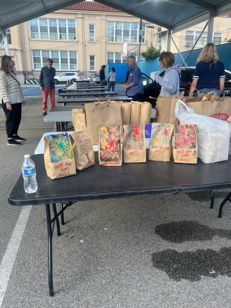 Table with paper bags decorated with colorful paintings, and a water bottle on a street under a canopy in a city setting.