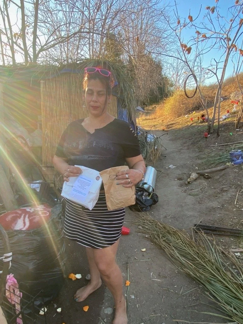 A woman with pink sunglasses and a black t-shirt stands outside on a dirt path, holding a paper bag and a white plastic bag, with a makeshift shelter and debris around her, during daylight with clear sky and bare trees in the background.