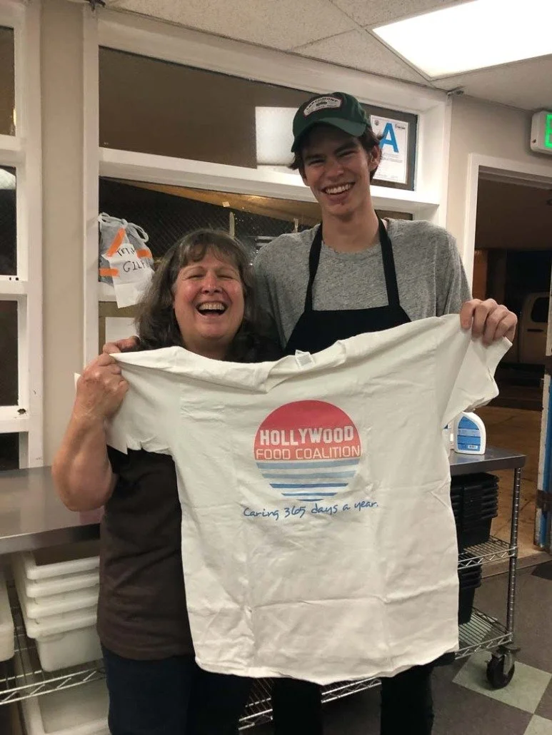 A woman and a young man smiling and holding a white T-shirt with the logo of Hollywood Food Coalition and the text 'Caring 365 days a year.' They are inside a room with shelves and a cart, indicating a food service or charity environment.