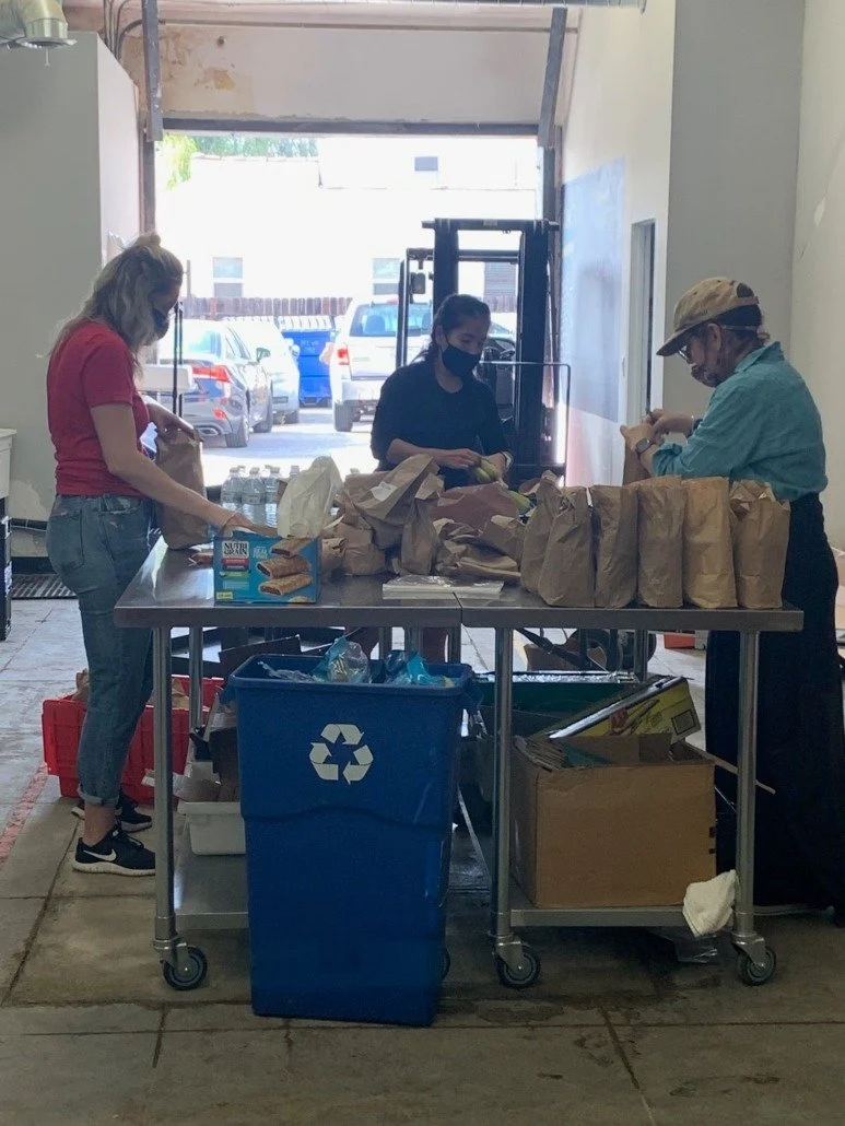 Three people packing or organizing brown paper bags on a metal table inside a garage or warehouse. There are water bottles and a box of Nutri-Grain bars on the table. A recycling bin and a cardboard box are underneath, and several parked cars are vis