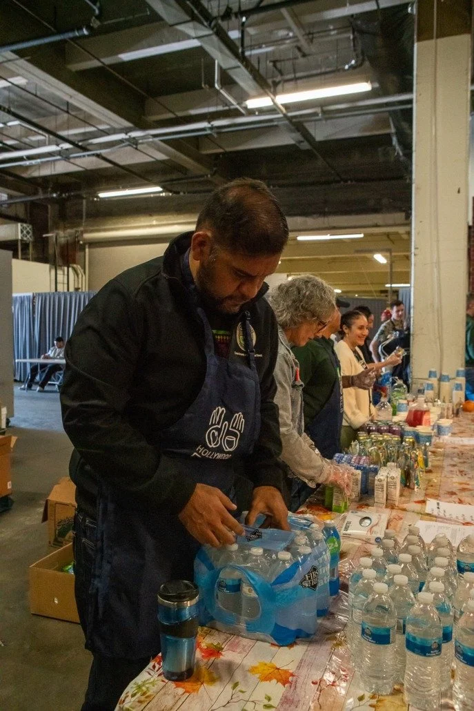 People volunteering at a food or supply distribution table in an indoor setting, with items like water bottles, snacks, and supplies.