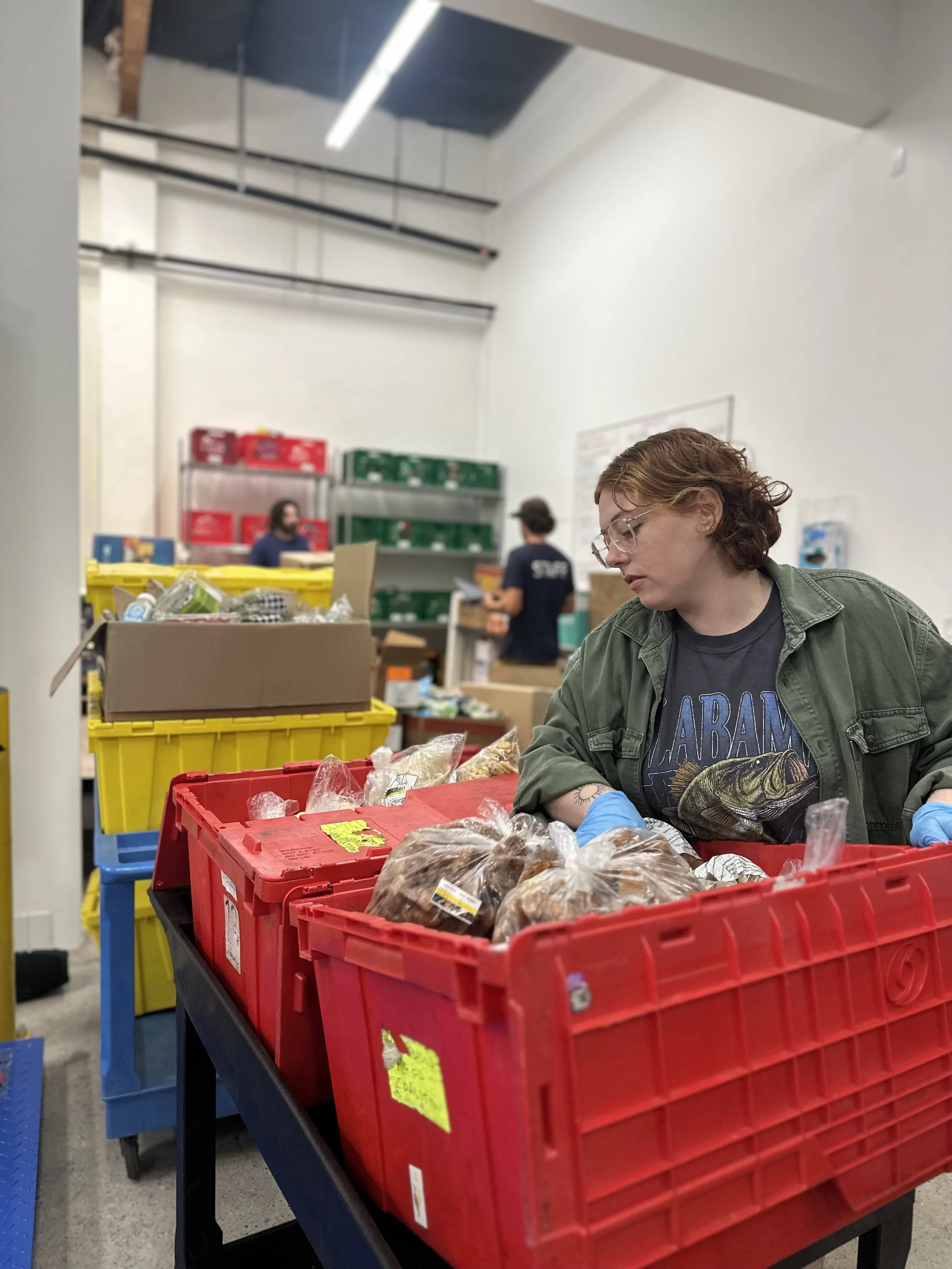 Person sorting packages of food in a warehouse or distribution center, with shelves and other workers in the background.