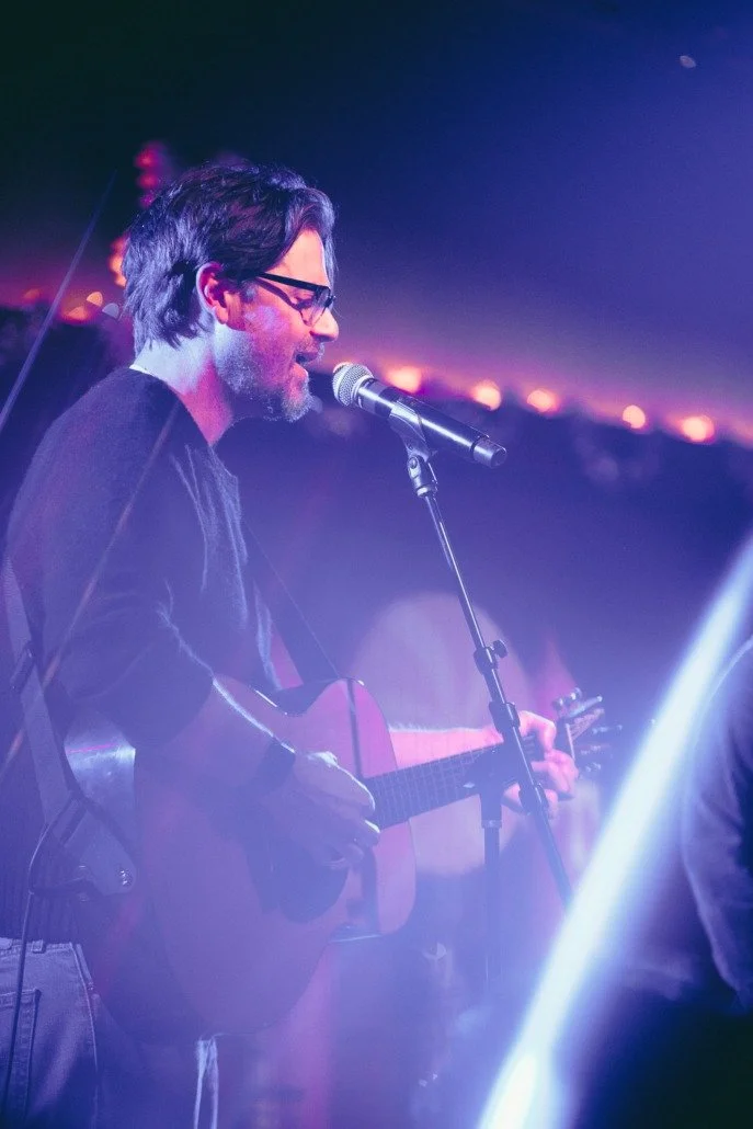 Man with glasses singing and playing guitar on stage at night, illuminated by purple and pink lights.