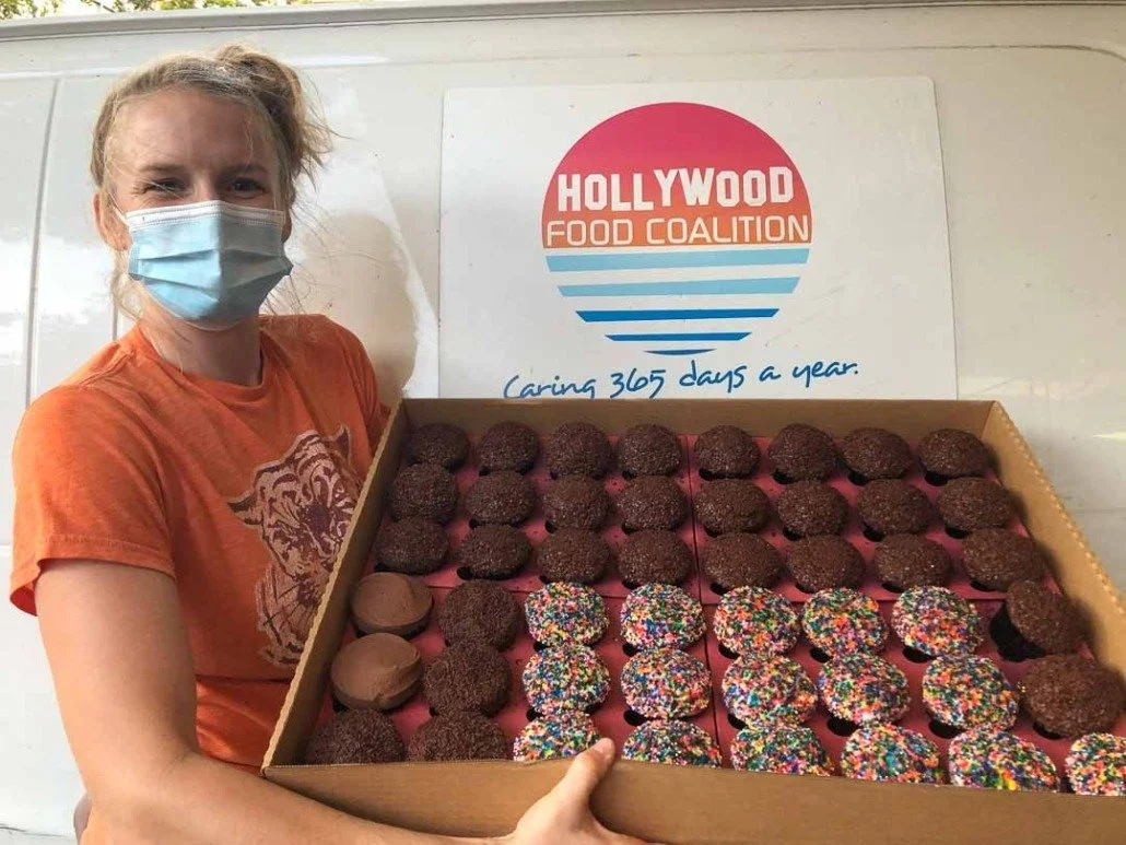 A woman wearing an orange T-shirt and face mask holding a large box of assorted cookies, including chocolate sprinkle and rainbow sprinkle cookies, in front of a sign that says 'Hollywood Food Coalition' and their slogan 'Caring 365 days a year.'