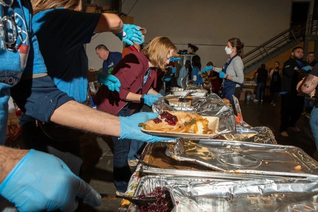 People serving and receiving food at a community meal event, with trays of food and people in masks and gloves.