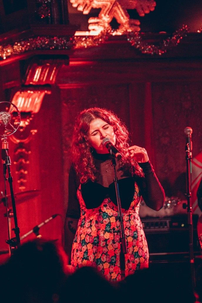 A woman with curly hair singing into a microphone during a live performance at a cozy, dimly lit venue with Christmas decorations.
