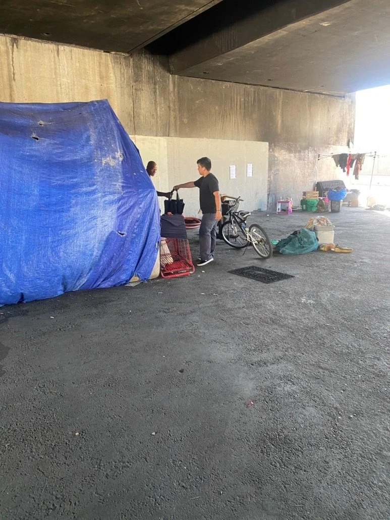 Two people under a bridge or overpass with makeshift shelter, surrounded by belongings and bicycles.