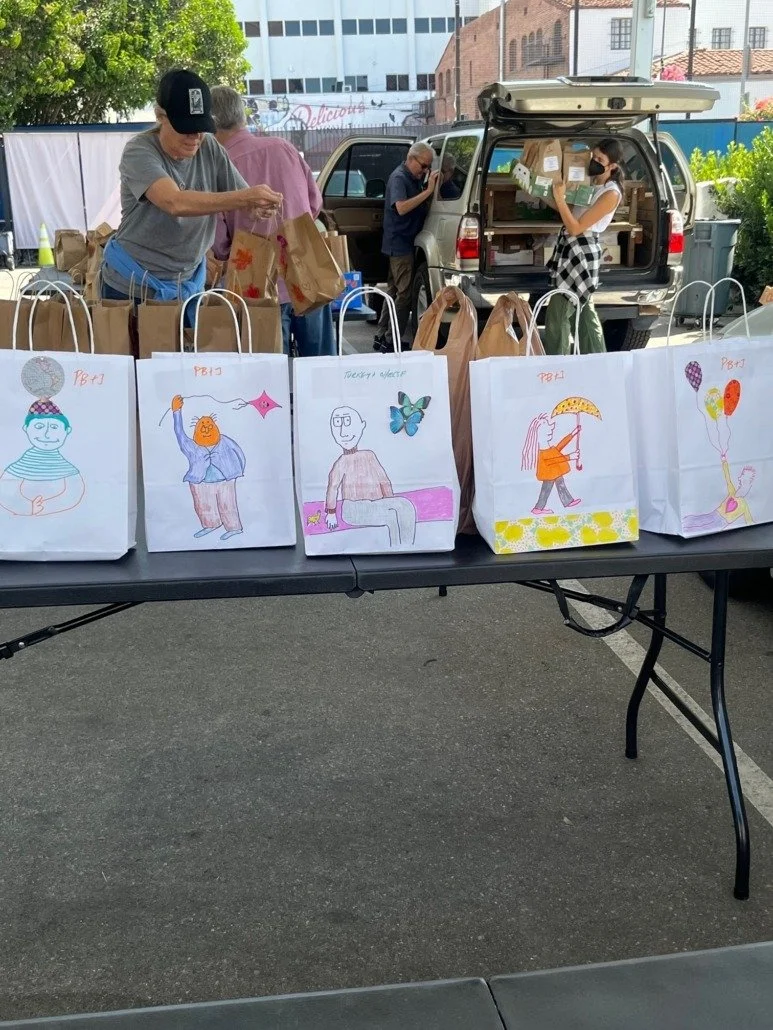 Table with colorful hand-drawn artwork and paper bags set up at an outdoor event, with people organizing items in the background.