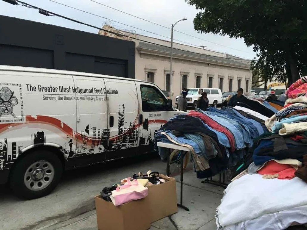 Street vendors selling clothes and other items outdoors next to a white van with the text "The Greater West Hollywood Food Coalition"