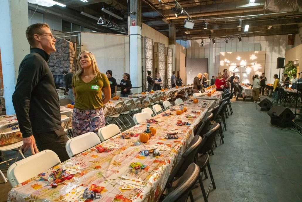 Indoor event space with long decorated tables, pumpkins, and candy, set for a celebration or gathering, with people socializing and a stage area in the background.
