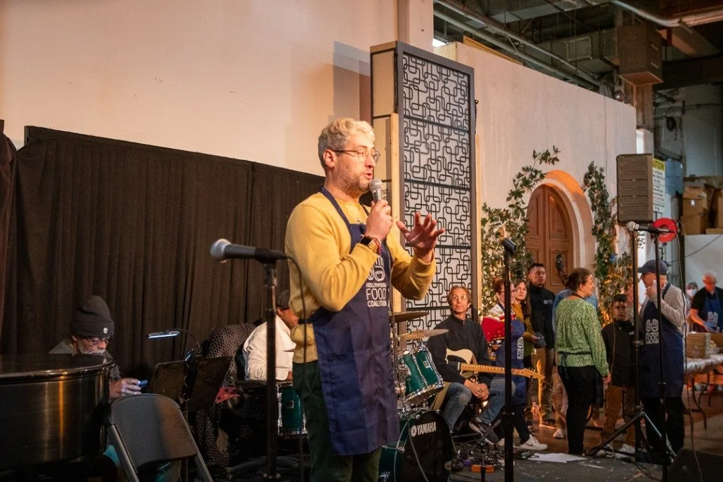 A man with glasses and gray hair holding a microphone, speaking on stage at an indoor event. He is wearing a yellow shirt and a dark apron. Behind him are musicians and people standing, with some plants and decorative elements in the background.