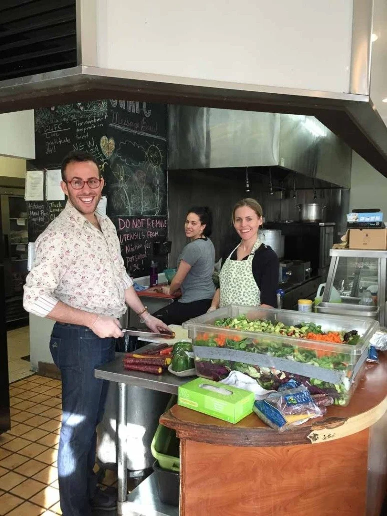 Three people preparing food in a commercial kitchen, with fresh vegetables on the counter, smiling.