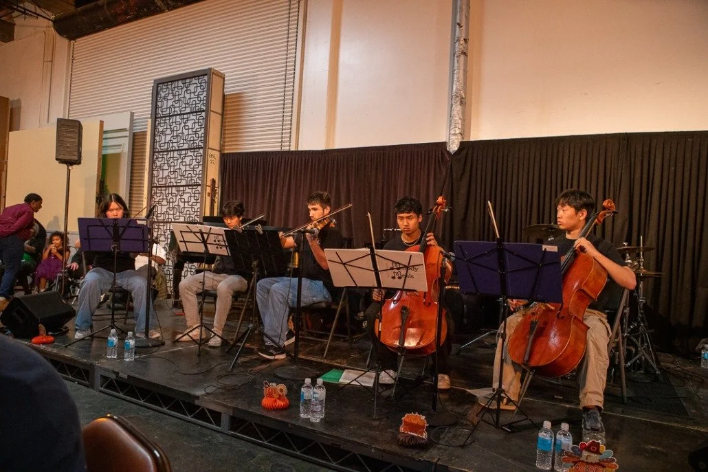 A group of five young musicians performing on stage with string instruments, including violin and cello, in a concert setting with black curtains and audience in the background.