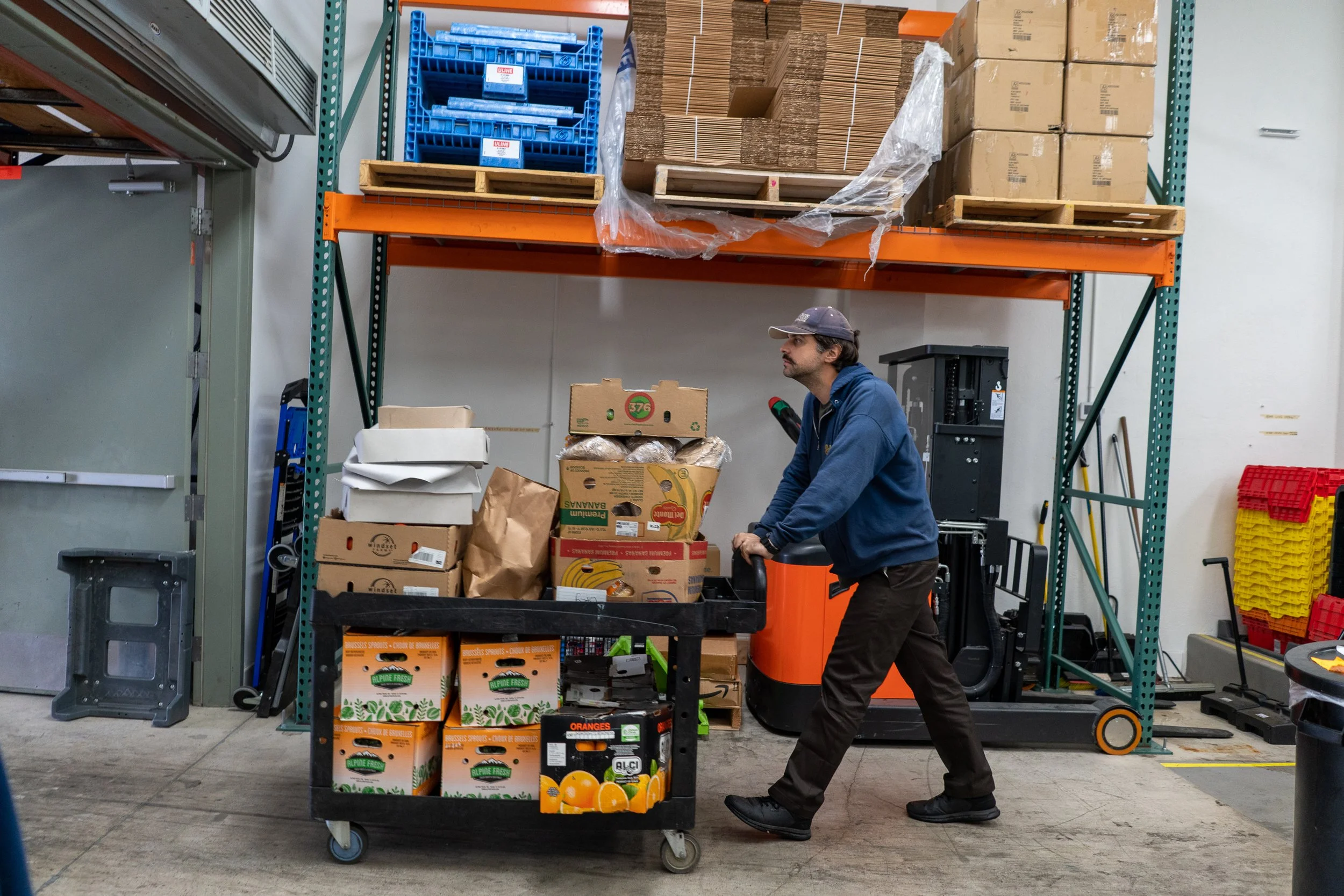 A man wearing a baseball cap and hoodie is pushing a cart filled with boxes of fruits and vegetables in a warehouse or storage room.